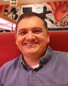 Fred R. DeLeon, smiling at camera, red background and Japanese writing on wall