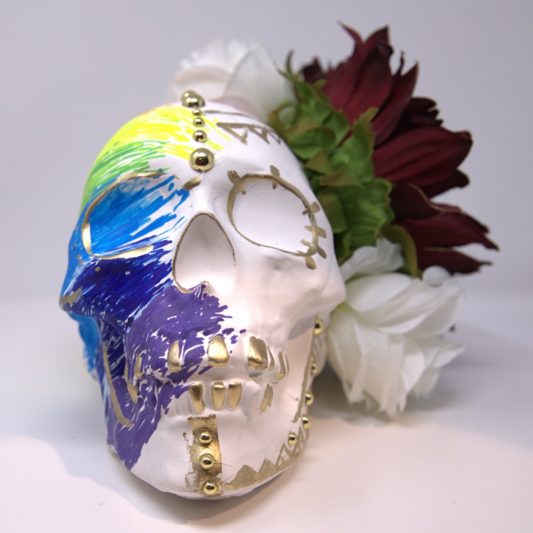 White skull with gold details and half painted in a rainbow pattern in representation of queerness, and with a side crown of flowers.